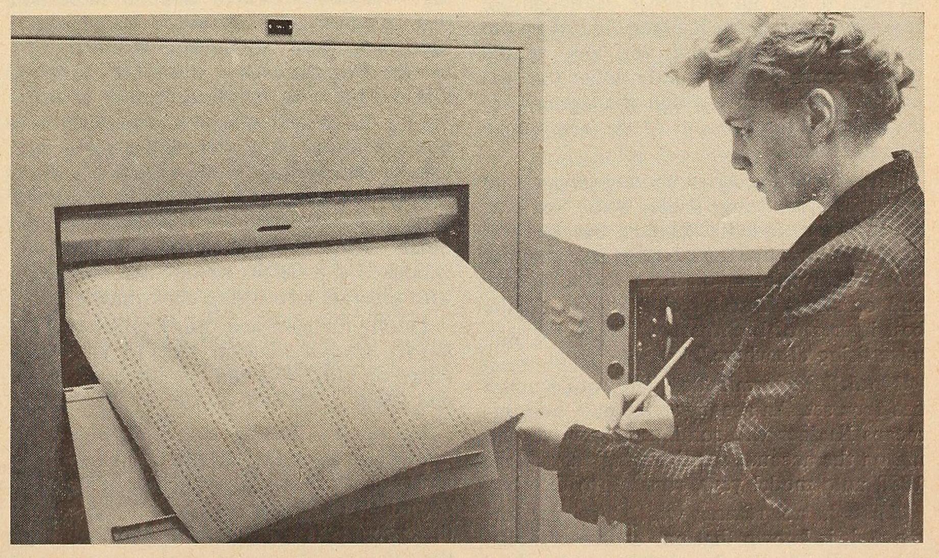 A black and white photograph of a woman standing at a large spool-feed printer. She is reading information off of a paper as it is being printed. The photo is scanned from a print magazine and appears slightly yellowed.