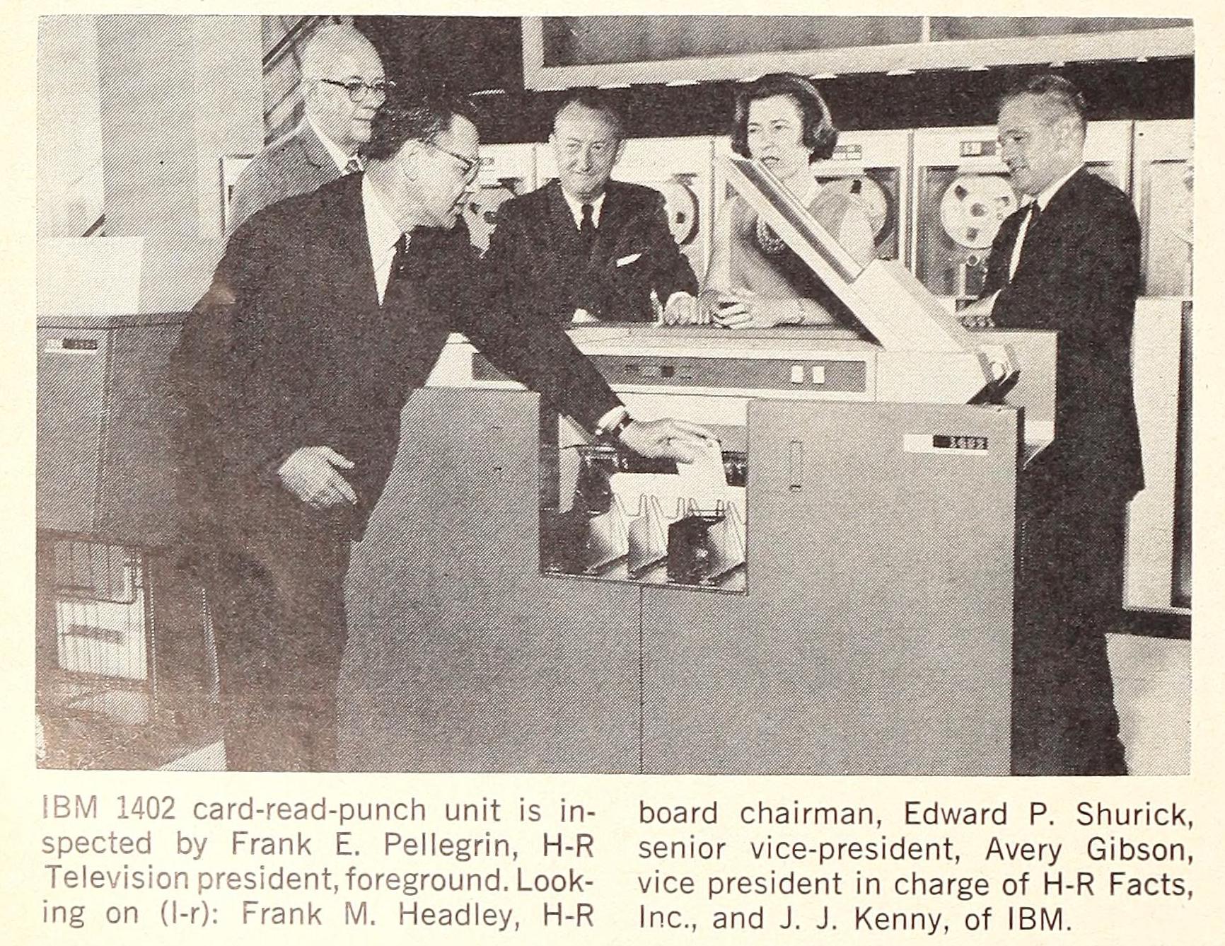 A black and white photo of men in suits and one woman standing around an IBM 1402 punch-card reader. There are large bays of magnetic tape in the backghround.